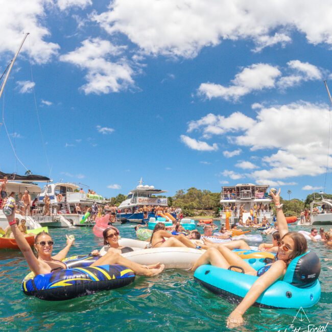 A group of people relax on colorful pool floats in the water, smiling and raising their arms, surrounded by boats under a bright blue sky with fluffy clouds. The scene is lively and festive.