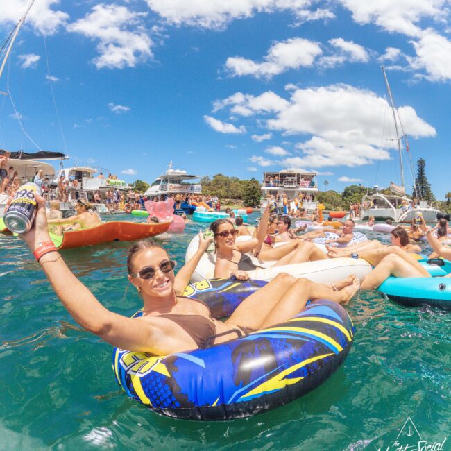 People relaxing on colorful inflatable tubes in clear blue water, holding drinks and smiling under a sunny sky, with boats and waterfront homes in the background. The scene is lively and festive.