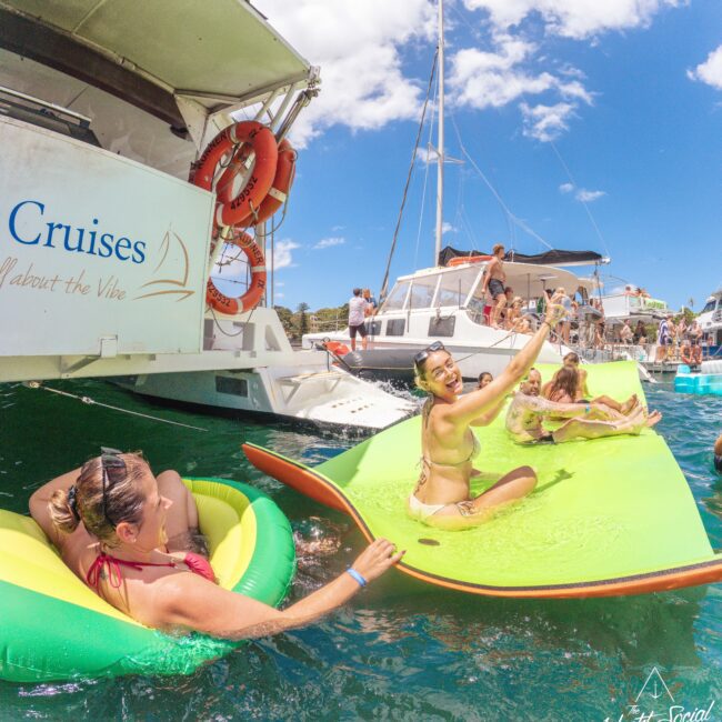 People relax and have fun on floats and a large mat in the water next to docked boats during a sunny day, with more people socializing on the boats. The scene is lively and festive under a bright blue sky.