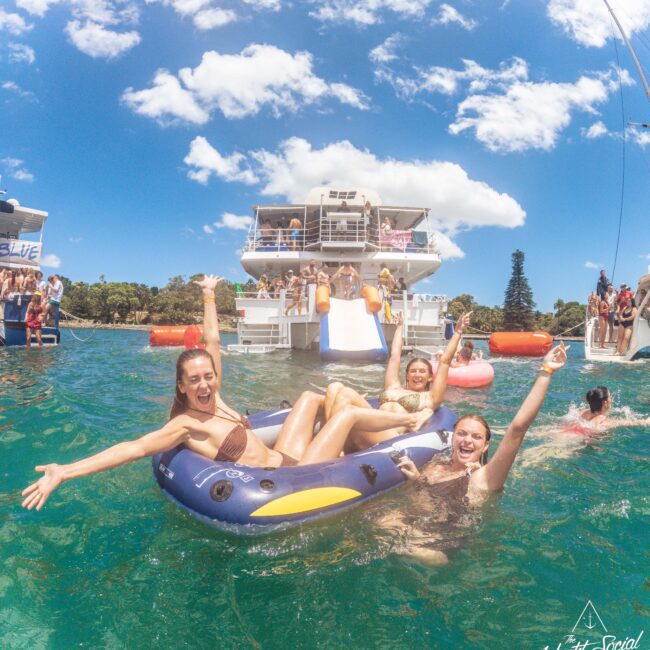 Three smiling women in swimsuits relax on an inflatable raft in the ocean, surrounded by people swimming and partying near yachts under a sunny, blue sky. The atmosphere is festive and lively.