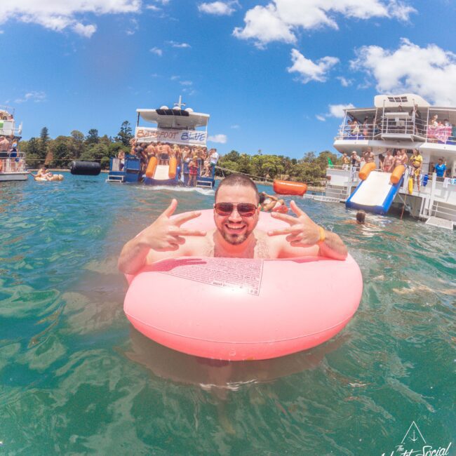 A smiling man in sunglasses floats on a pink inner tube in a lake, flashing peace signs. Boats and people enjoying water activities are in the background under a sunny, blue sky.