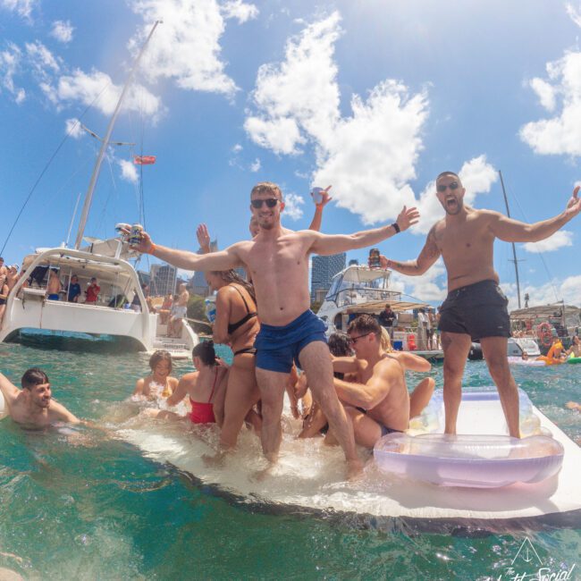 A group of people in swimwear enjoy a lively yacht party on the water, standing and sitting on floating mats near boats, with blue skies and city buildings in the background.