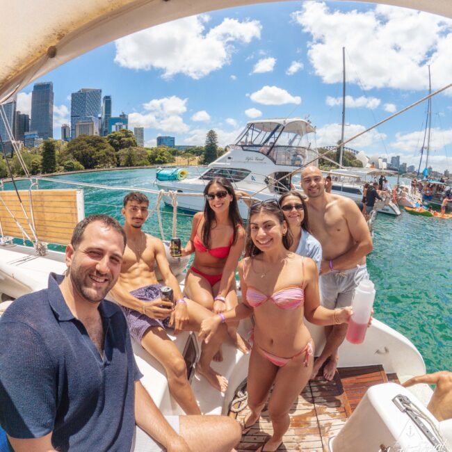 A group of six smiling young adults in swimsuits enjoy a sunny day on a boat, with city buildings and other boats visible in the background on calm blue water.