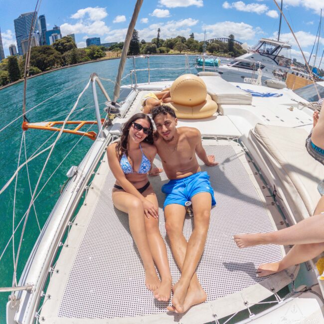 A smiling couple in swimwear relaxes on the netting of a catamaran, with turquoise water and a city skyline in the background under a bright blue sky. Another person's legs are partially visible in the frame.
