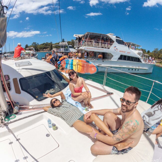 Three people relax and smile on the deck of a sailboat under a sunny sky, with other boats and people visible on the water in the background.