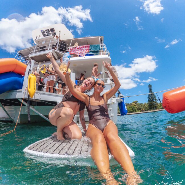 Two women in swimsuits smile and pose on a floating mat in the water near a yacht, with other people onboard. The sky is blue with white clouds, and the scene is lively and festive.