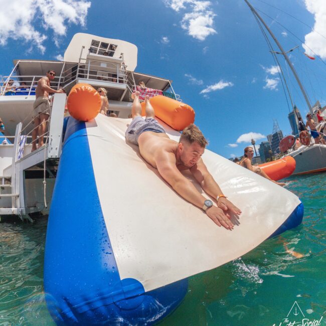 A man slides down an inflatable water slide from a yacht into turquoise water, surrounded by people enjoying a sunny day. Other boats and blue sky with clouds are visible in the background.