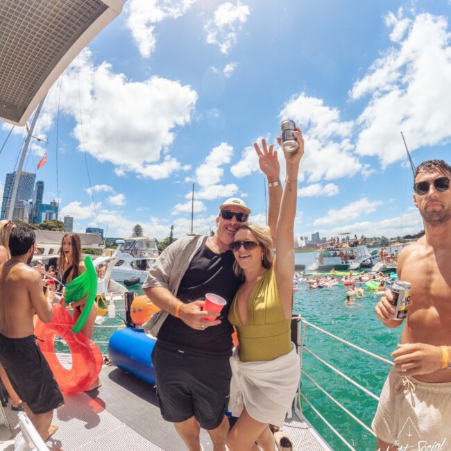 Two people smile and pose with drinks on a boat during a sunny day, surrounded by other boats and people enjoying a lively party on the water. The sky is blue with scattered clouds.