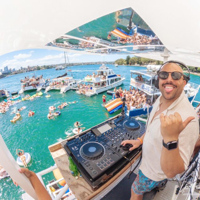 A DJ wearing sunglasses and headphones smiles and gestures to the camera while playing music on a boat. In the background, people party on boats and floaties in the water under a sunny sky.