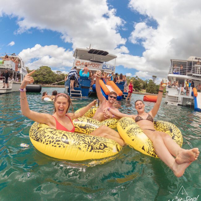 Three smiling people relax on inflatable rafts in the water, making peace signs, with boats and slides in the background under a partly cloudy sky. The scene is lively and joyful at a social event.