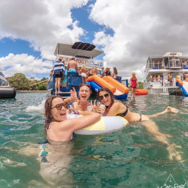 Three women in swimsuits smile and relax on a pool float in the water, surrounded by people, boats, and water slides, under a cloudy sky at a lively social event.