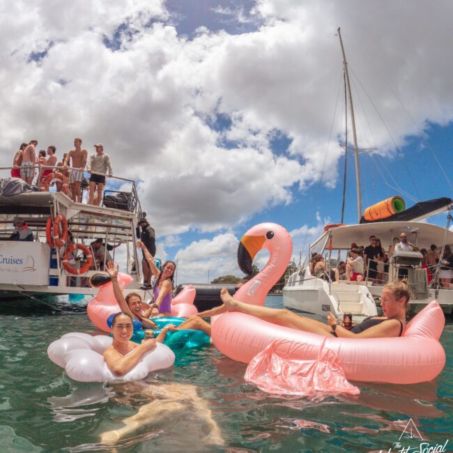People relax and have fun on inflatable pool floats shaped like a flamingo and a cloud in the water near two boats, under a partly cloudy sky. Others stand and socialize on the boats in the background.