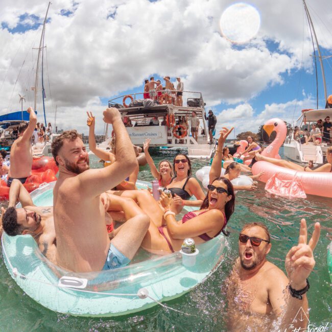 A group of people in swimsuits enjoy a lively party on inflatable floats in the ocean near boats, laughing, holding drinks, and making peace signs under a cloudy sky.