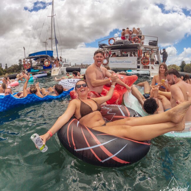 A group of young adults relax on inflatable floats in the water near boats, laughing, holding drinks, and enjoying a sunny day with partly cloudy skies. Some people are on the boats in the background.