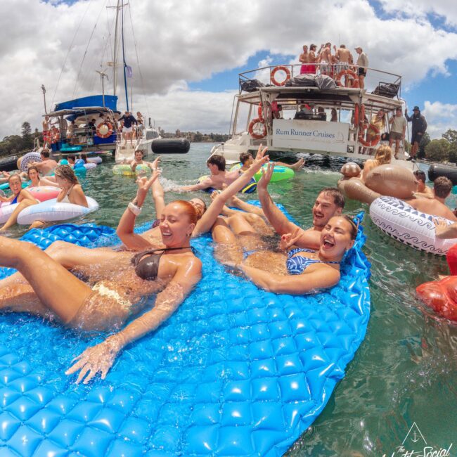 Three women laugh and splash on a blue inflatable raft in the water, surrounded by people on floaties and boats, enjoying a lively daytime party under a partly cloudy sky.