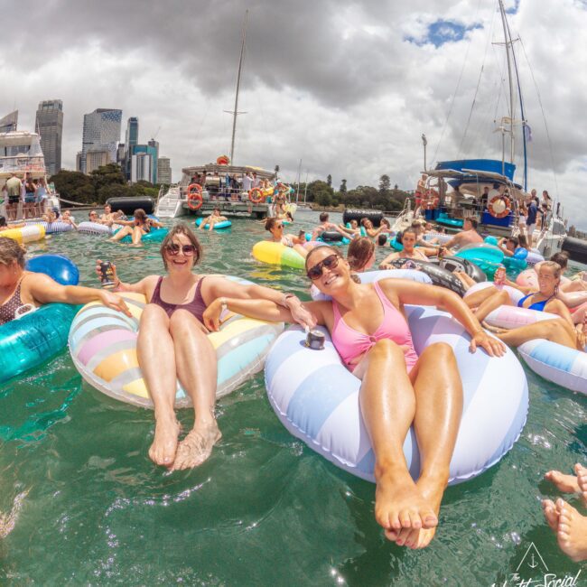 Two women in sunglasses relax on inflatable tubes in the water, surrounded by other people on floats. Boats and a cityscape are visible in the background under a cloudy sky.