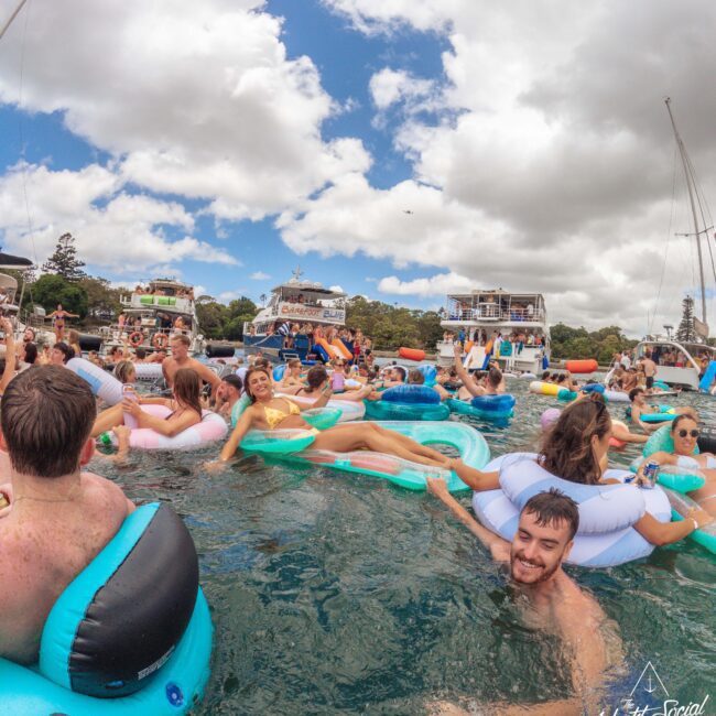 A large group of people relaxing on inflatable floats in the water near several docked boats, enjoying a sunny day under partly cloudy skies. Some people are smiling at the camera. The atmosphere is lively and festive.