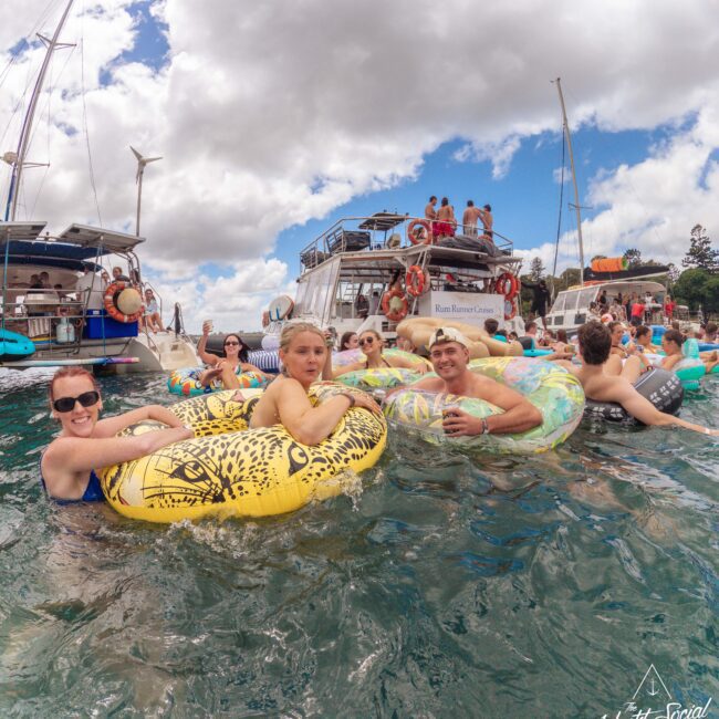 A group of people relax on colorful inflatables in the water near several boats, with others socializing on the decks under a partly cloudy sky. The scene looks lively and summery.