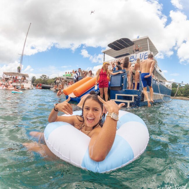 A smiling woman in a striped inner tube floats in turquoise water, making a shaka hand gesture. Behind her, a group of people enjoy a party on a boat under a bright, partly cloudy sky.