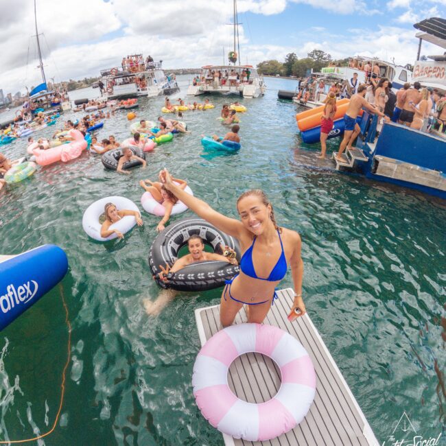 A woman in a blue bikini smiles and flashes a peace sign on a dock surrounded by people in colorful floaties at a lively pool party on the water, with boats and a crowd in the background under a partly cloudy sky.