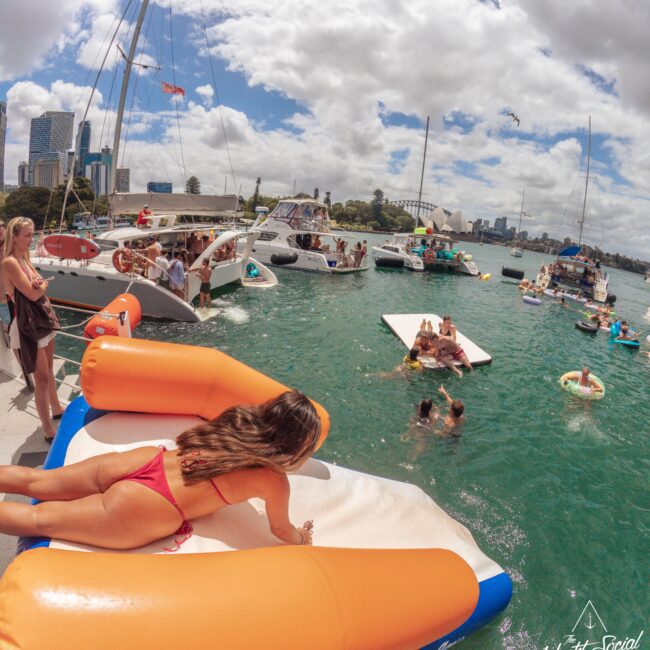 A woman in a red bikini lounges on a large inflatable pizza slice in the water near several anchored boats. People are swimming and relaxing, with a city skyline and cloudy sky in the background.