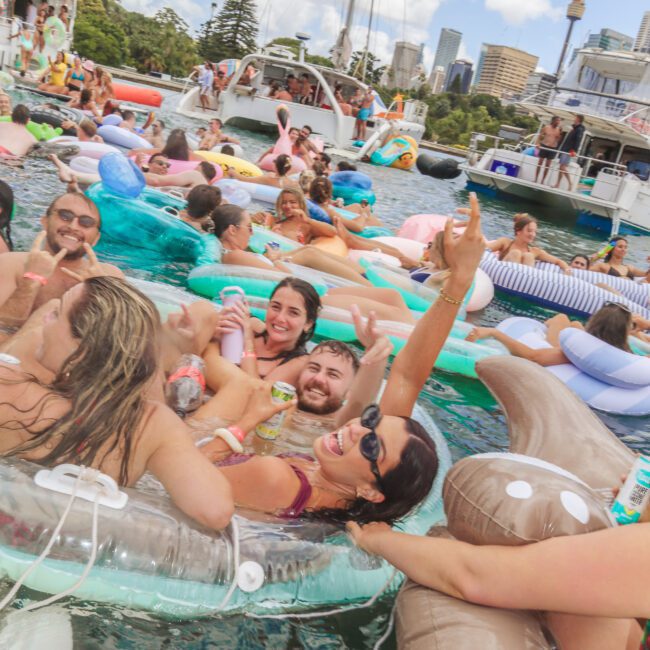 A group of people relax and smile on colorful pool floats in the water at a lively outdoor party, with boats and a city skyline visible in the background.