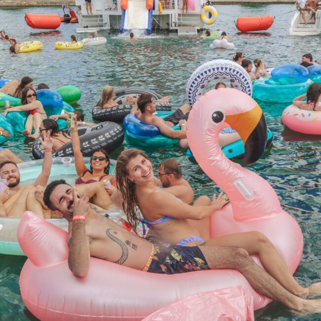 A group of people float and relax on colorful inflatable pool floats in the water during a lively summer party. Some are smiling and waving, while others lounge and enjoy the festive atmosphere.