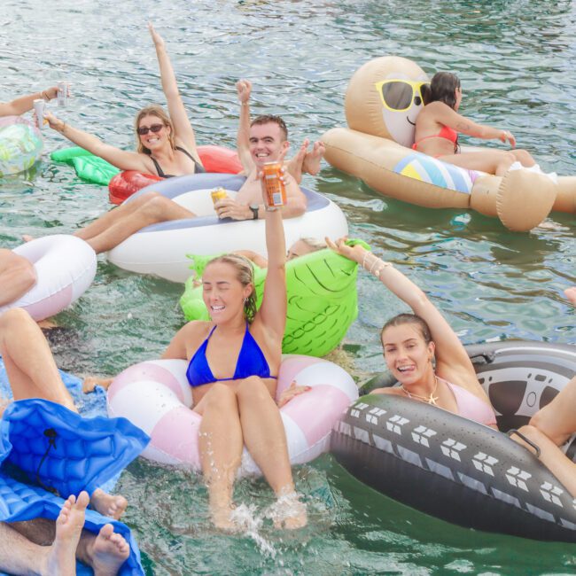 A group of people in swimsuits relax and have fun on colorful inflatable floats in the water, smiling, raising their arms, and holding drinks on a sunny day.