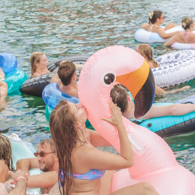 A group of people relax and have fun floating on colorful inflatable tubes and pool floats, including a large pink flamingo, in a body of water on a sunny day.