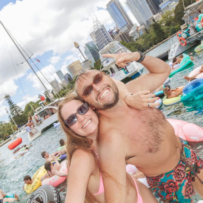 A smiling man and woman take a selfie while standing in the water at a pool party, surrounded by people on colorful inflatables. City skyscrapers and trees are visible in the background under a partly cloudy sky.