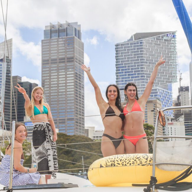 Four women in swimsuits stand and sit on a sailboat, smiling and waving toward the camera. Tall city buildings and trees are visible in the background under a partly cloudy sky.