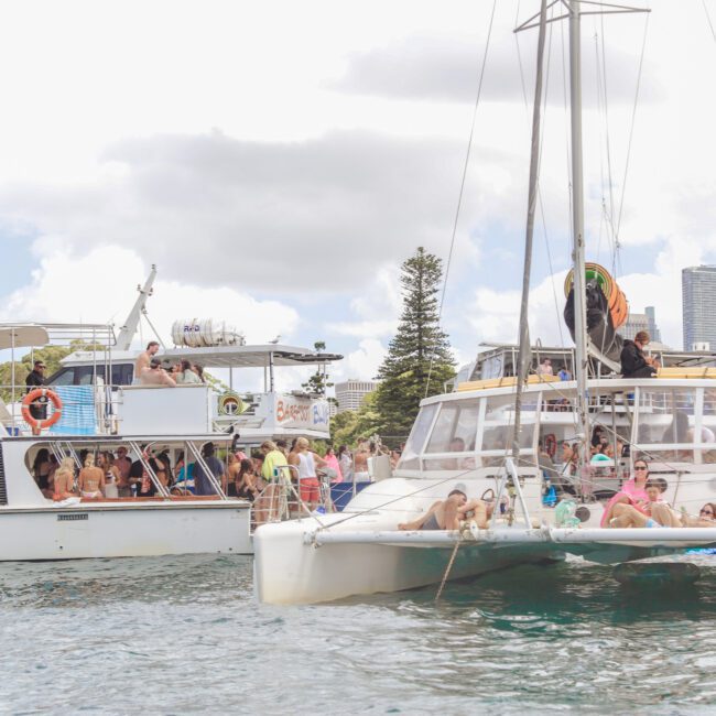 Two boats filled with people relaxing and socializing are anchored side by side on the water near a city skyline; some people are sunbathing while others stand and enjoy the view.
