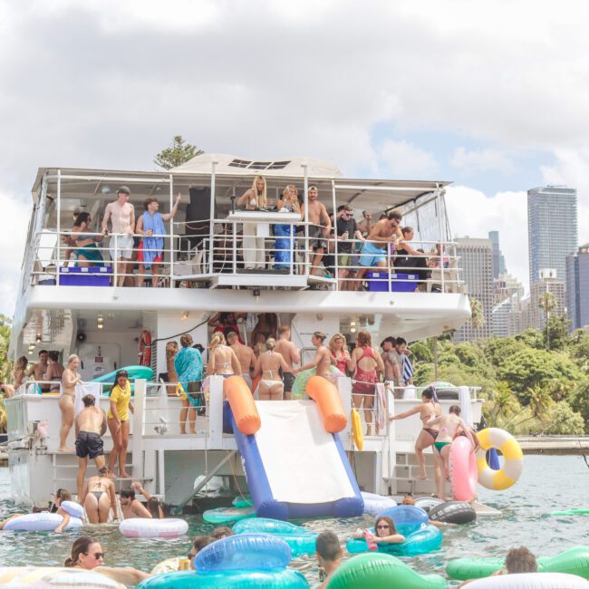 A large group of people enjoy a party on a double-decker boat with inflatable pool floats in the water. The boat is near a city with tall buildings and surrounded by greenery under a partly cloudy sky.