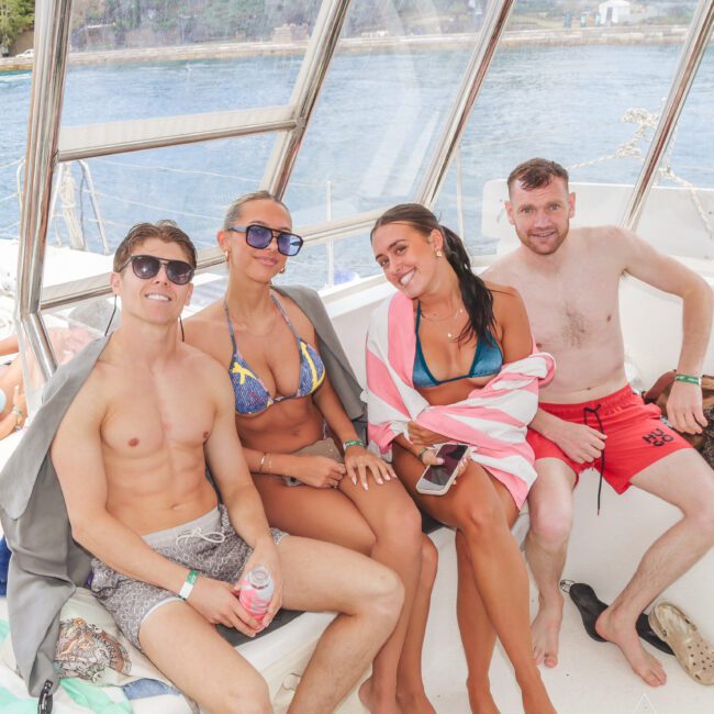 Four people sit close together on a boat, smiling at the camera. Two women in bikinis are in the middle, one wrapped in a towel. Two men in swim trunks are on either end. The ocean and coastline are visible through the windows.
