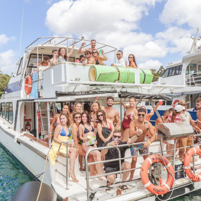 A large group of people in swimsuits pose and smile on a white yacht docked by the water, with other boats and trees in the background on a sunny day.