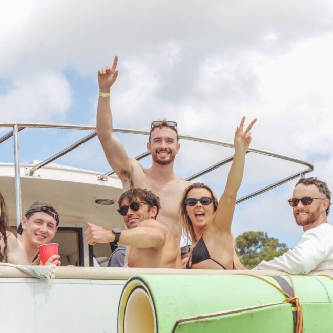 A group of seven young adults smile, cheer, and pose on a boat on a sunny day. Some wear sunglasses and swimsuits, and one holds up a peace sign. Surfboards are visible in the foreground.