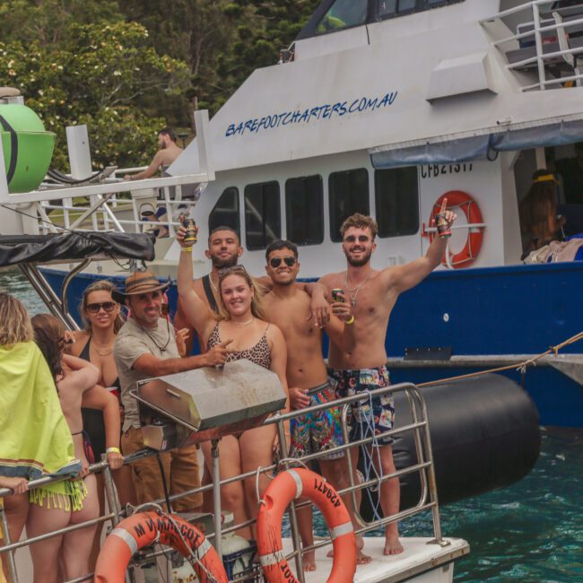 A group of people in swimwear smile and pose on a boat, holding drinks and enjoying a tropical day. Another boat is anchored nearby, and the scene is lively, with greenery in the background.