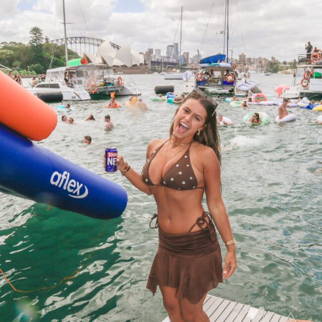 A smiling woman in a brown bikini and skirt stands on a dock holding a drink, with boats and people swimming and floating in the water behind her. The Sydney Opera House and city skyline are visible in the background.