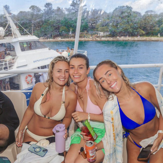 Three women in swimsuits smile and pose together on a boat, with water and trees visible in the background. They appear relaxed and are holding drinks, enjoying a day by the water.