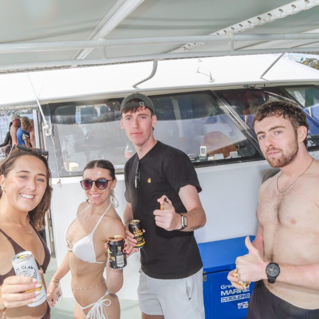 Four young adults stand on a boat deck, smiling and holding drinks. Two women wear swimsuits and sunglasses; two men wear swim trunks, one shirtless and one in a t-shirt. Water and other boats are visible in the background.