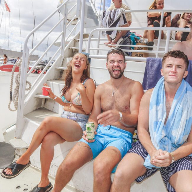 Three people in swimsuits sit on a boat, laughing and smiling, with one wrapped in a striped towel. Other people are in the background, and the atmosphere is lively and sunny.