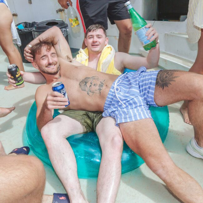 Two men in swimwear relax on a pool float, smiling and holding drinks at a lively gathering on a sunny boat deck, surrounded by other people in swimwear.