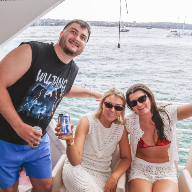 Three people smile and pose on a boat, holding drinks. The man wears a black Wu-Tang shirt and shorts; the two women, in summer attire, sit and wave. Water and boats are visible in the background.