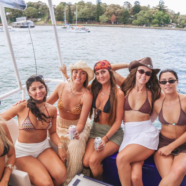Six women in swimsuits and hats sit and smile on a boat by the water, holding drinks. Trees and other boats are visible in the background on a sunny day.