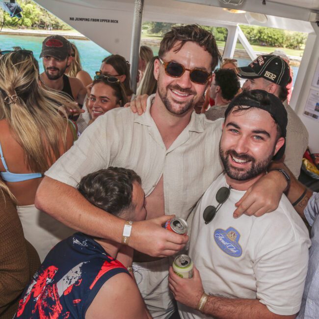 A group of people smiling and enjoying drinks at a lively boat party. Two men stand in the center, arms around each other, while others mingle in swimwear and summer clothes. Sunlight streams in from the water outside.