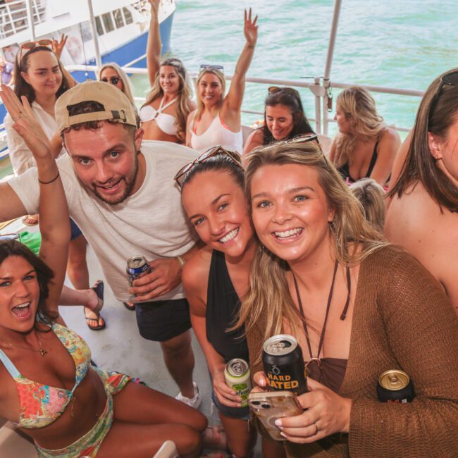 A group of young adults smiling and posing with drinks on a crowded party boat, with blue water and another boat visible in the background. Some people are waving, and everyone appears to be having fun.