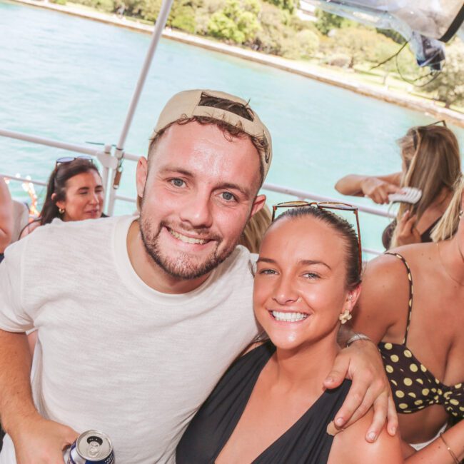 A group of young adults smiling and enjoying drinks on a boat. The man in front has his arm around a woman, and others in the background are laughing. Sunny weather and water can be seen behind them.