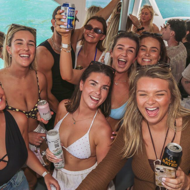 A group of young adults in swimwear smile and hold drinks while gathered closely together on a boat, enjoying a sunny day by the water. Other people mingle in the background.