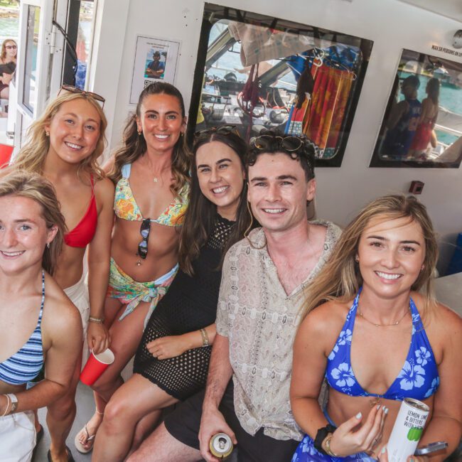 A group of six smiling young adults, dressed in swimwear and casual clothes, pose together on a boat. Some are holding drinks, and bright sunlight streams in from outside, with water and other people visible in the background.