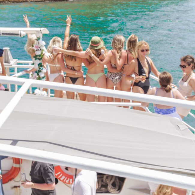 A group of women in swimsuits stand together on a boat, facing the water with their arms around each other, enjoying a sunny day. Other people onboard relax nearby. The sea is turquoise and calm.